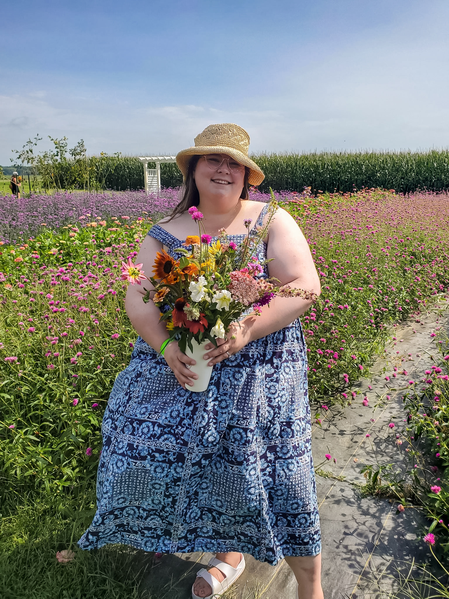 Photo of Nicole Loe with a bouquet of fresh cut flowers in a field. She is wearing a blue dress and a cute hate.