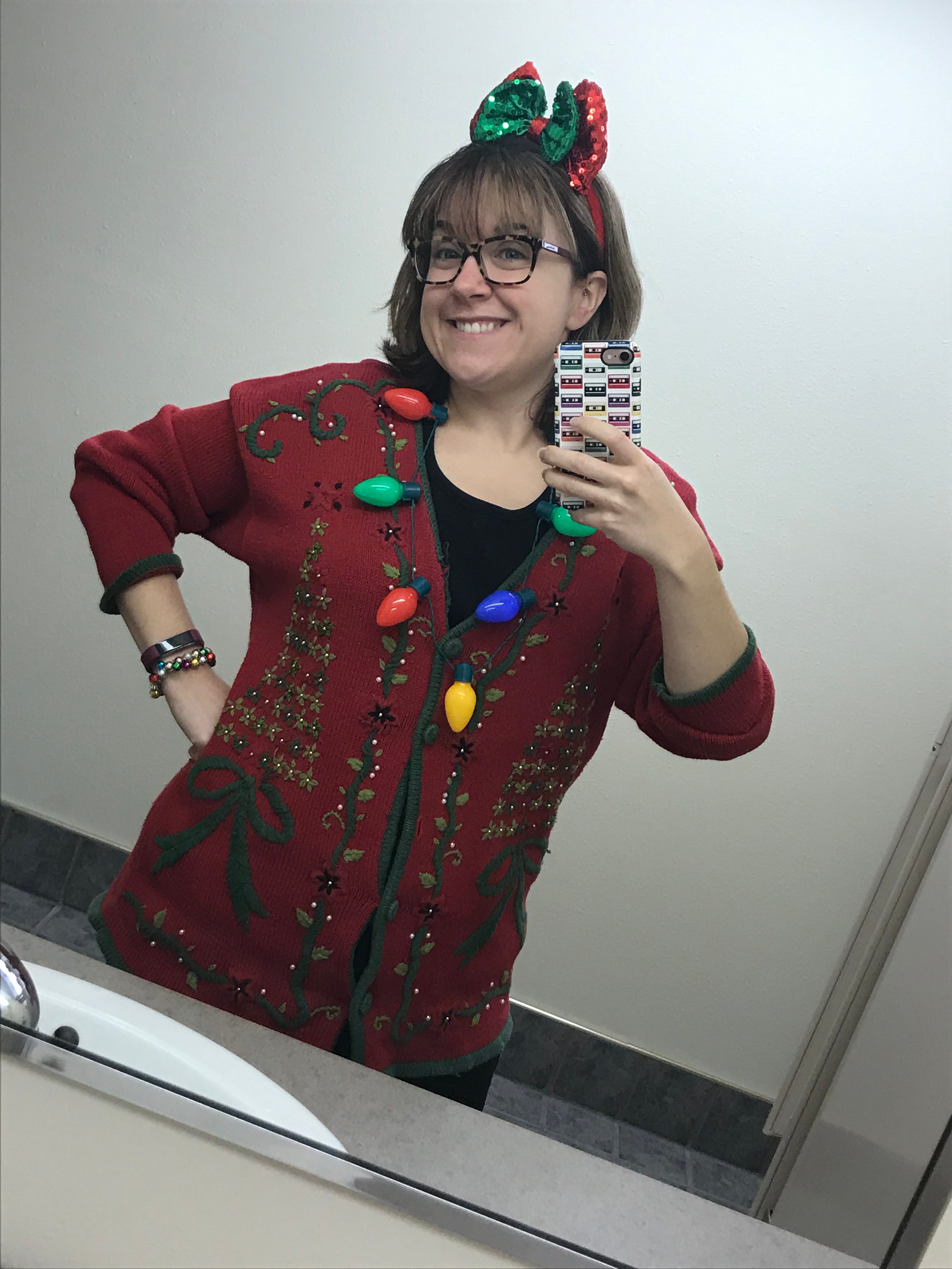 Natasha in a bathroom selfie wearing an Ugly Christmas sweater. It is red with sewn on Christmas trees and a festive bow headband.